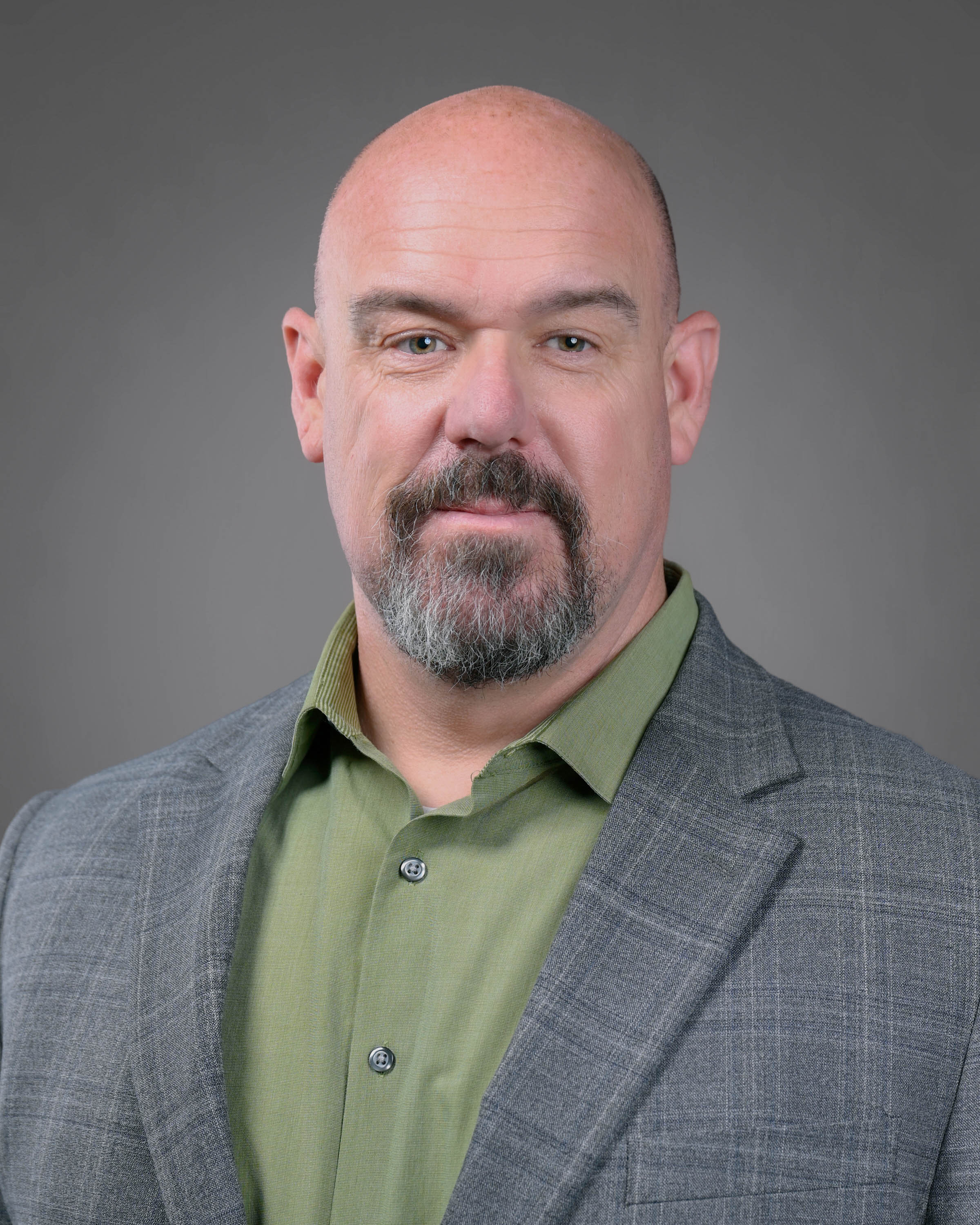 Man with a goatee and shaved head wearing a grey jacket and green shirt